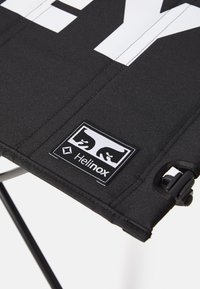 Zwarte stoffen stoel met een witte grafische print en een Helinox-logo patch. Heeft een duurzame structuur en een vouwmechanisme voor draagbaarheid.