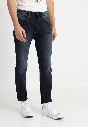 Mand iført mørkeblå slim-fit jeans med optrukne manchetter, hvide sneakers og hvide sokker mod en ensfarvet lys baggrund.