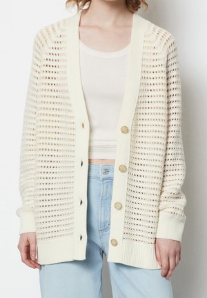Femme portant un cardigan crème en maille ouverte boutonné sur un crop top blanc et un jean taille haute bleu clair, debout devant un fond blanc.