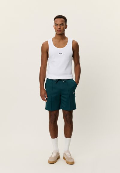 Débardeur blanc sans manches avec un petit logo noir, associé à un short turquoise avec poches avant et taille à cordon. Baskets beige.