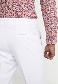 Pantalones blancos con una textura suave, que cuentan con un bolsillo trasero con cierre de botón. Una camisa de patrón floral colorido es parcialmente visible.