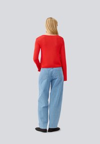 Haut rouge à manches longues à côtes, associé à un jean bleu clair à coupe décontractée, vu de dos. Texture douce, design décontracté. Chaussures noires.