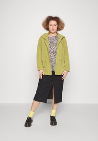 Giacca in suede verde, top con stampa leopardata, gonna nera con spacco laterale, calze gialle e scarpe nere. Sfondo semplice, vista frontale.