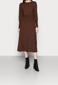 Brun långärmad klänning med kroppsnära midja och utsvängd kjol. Slät tyg med matt yta. Svarta klackstövlar med blockklack bärs.