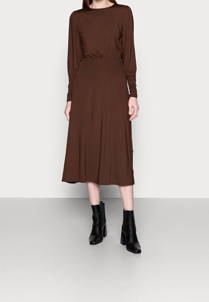 Braunes Langarmkleid mit taillierter Oberteile und ausgestellt Rock. Glattes Material mit matter Oberfläche. Schwarze Blockabsatz-Stiefeletten getragen.