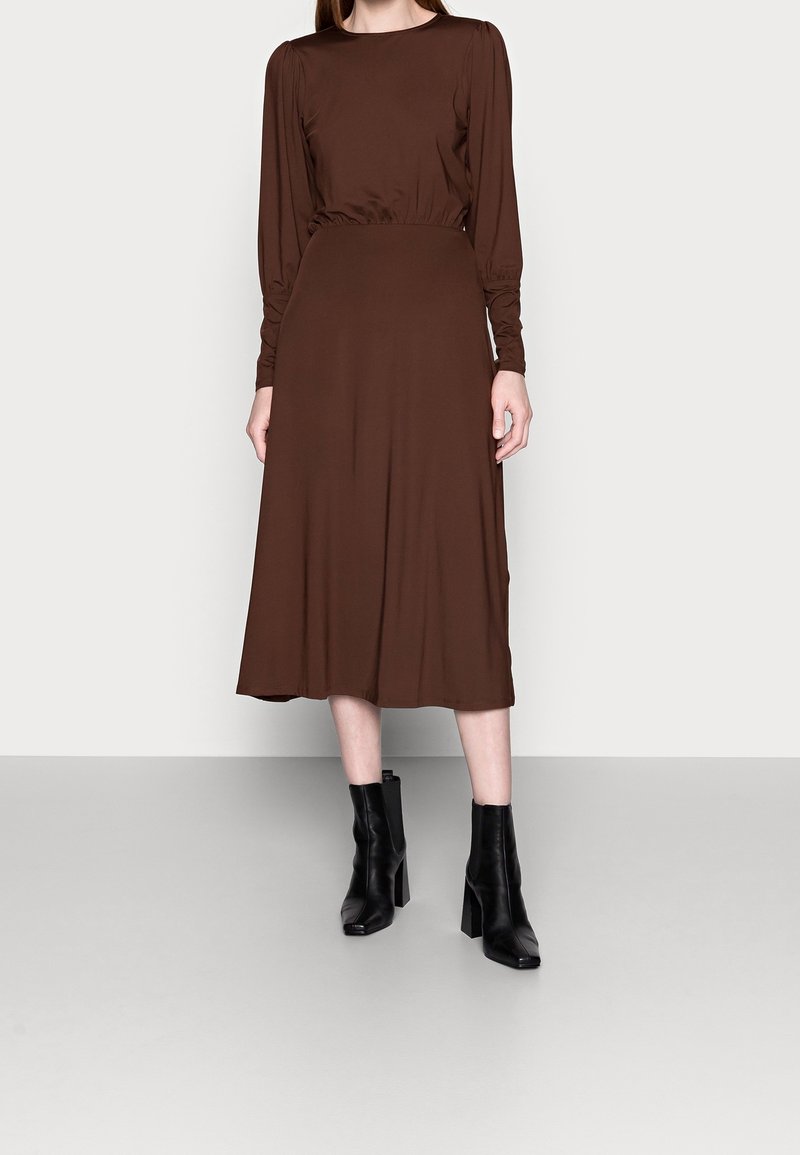 Brun långärmad klänning med kroppsnära midja och utsvängd kjol. Slät tyg med matt yta. Svarta klackstövlar med blockklack bärs.