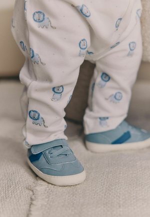 Babyhosen aus weißer Baumwolle mit blauen Löwendrucken, kombiniert mit blauen Wildledersneakern mit weißer Gummisohle und Klettverschluss.