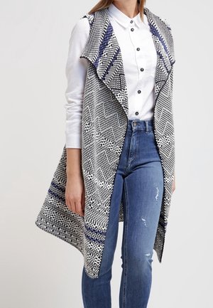Gilet à motifs gris et blanc avec un col drapé, des poches latérales et des accents bleus contrastants, porté sur une chemise blanche et un jean bleu.