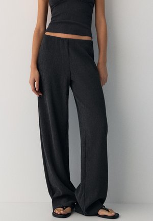 Vrouw draagt zwarte wide-leg broek met textuur en een bijpassende mouwloze crop top, staand tegen een lichtgrijze muur.