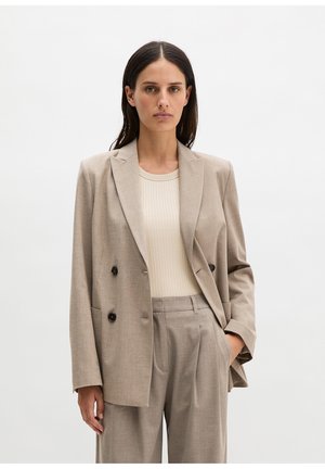 Frau trägt beige Zweireiher-Blazer und passende weite Hosen, kombiniert mit einem cremefarbenen Ripptop, steht vor einem schlichten Hintergrund.