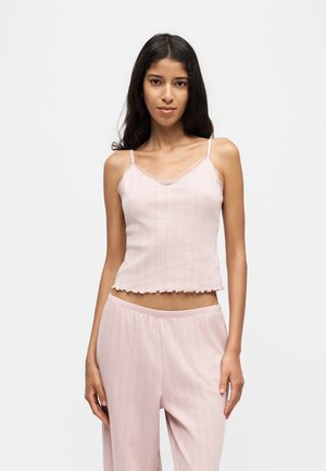 Jeune femme aux longs cheveux foncés portant un débardeur côtelé rose clair et un pantalon large assorti sur un fond uni.