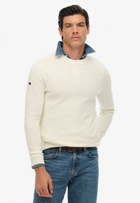 Biały sweter z teksturowanej bawełny, z okrągłym dekoltem, noszony na niebieskiej koszuli z kołnierzykiem. Posiada długie rękawy i subtelne logo na rękawie.