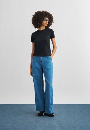 Mujer con cabello rizado que lleva una camiseta negra de manga corta, jeans azules de pierna ancha, zapatos negros y gafas de sol, de pie con una mano en el bolsillo.