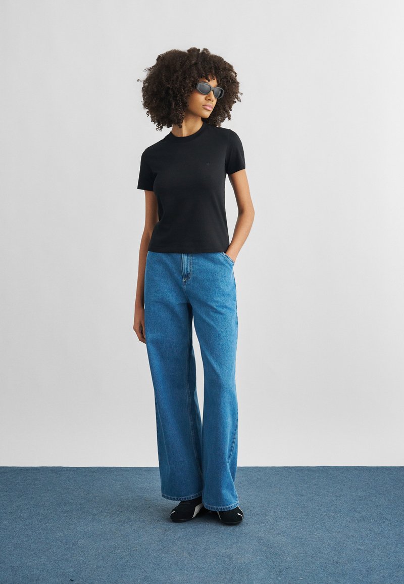 Woman with curly hair wearing black short-sleeve top, wide-leg blue jeans, black shoes, and sunglasses, standing with one hand in pocket.