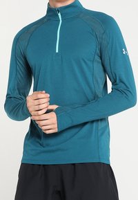 Teal langmouwige sporttop met een half-rits kraag en een contrasterende lichtblauwe rits. Heeft een gestructureerde stof en een aansluitend ontwerp.