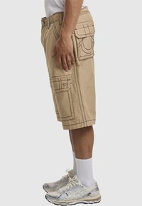 Beige Cargo-Shorts mit aufgesetzten Taschen, kontrastierendem Nähen und lässigem Schnitt. Getragen mit grauen Sneakers und weißen Crew-Socken.