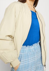 Giubbotto bomber beige con zip frontale, polsini a coste e una texture liscia, indossato sopra una maglia blu e pantaloni a quadri azzurri chiari.