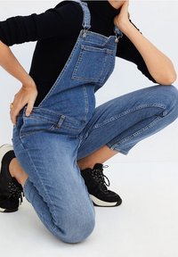 Denim overalls with adjustable straps, front pocket, and tapered legs. Worn with black sneakers, featuring a ribbed black top.