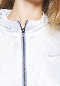 Bílá síťovaná zipová bunda s šedým zipem a fialovými akcenty. Má kapuci a malé logo Nike na hrudi.