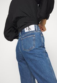 Blue denim jeans with a high waist, featuring two back pockets and a white label with "Calvin Klein Jeans" on the waistband.