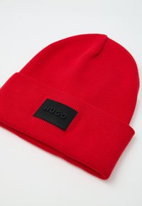 Rød strikket hue med foldet manchet. Har et sort rektangulært mærke med prægede bogstaver "HUGO." Stoffet virker blødt og tekstureret.
