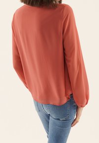 Blusa corallo con maniche lunghe e svolazzanti e una vestibilità ampia, realizzata in tessuto leggero. L'orlo è leggermente più lungo sul retro. Abbinata a jeans blu.