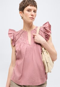 Γυναίκα με κοντά μαλλιά που φοράει ροζ μπλούζα με βολάν στα μανίκια και κρατάει μια μπεζ τσάντα ώμου μπροστά σε μονόχρωμο φόντο.