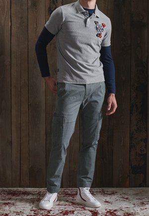 Mężczyzna w szarej koszulce polo na granatowej koszuli z długim rękawem, szare spodnie bojówki i białe wysokie trampki, stojący na wzorzystym dywanie.