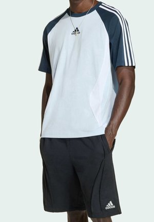 Mężczyzna w białej i granatowej koszulce Adidas oraz czarnych szortach Adidas, stojący z jedną ręką w kieszeni na jednolitym tle.