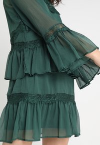 Grön chiffongklänning med lager av volanger, spetsdetaljer och trumpetärmar. Tyget har en mjuk, genomskinlig textur med en flödande design.