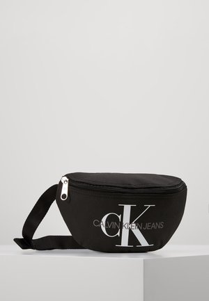 Schwarze Calvin Klein Jeans Bauchtasche mit weißem Logo, Reißverschluss und verstellbarem Tragegurt auf weißer Oberfläche.