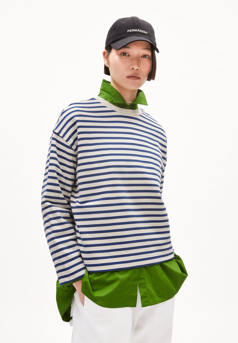 Maglia a righe con maniche lunghe in blu navy e bianco, sovrapposta a una camicia colletto verde brillante, abbinata a pantaloni bianchi e un cappellino nero.