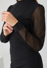 Black dress featuring long sleeves made of sheer fabric with polka dot pattern. High neckline and fitted body with textured sleeve accents.
