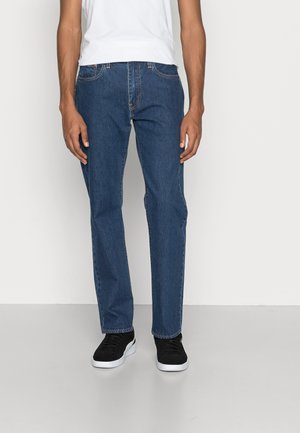 Hombre vistiendo jeans ajustados de color azul, zapatillas negras con suelas blancas y una camiseta sin mangas blanca frente a un fondo liso.