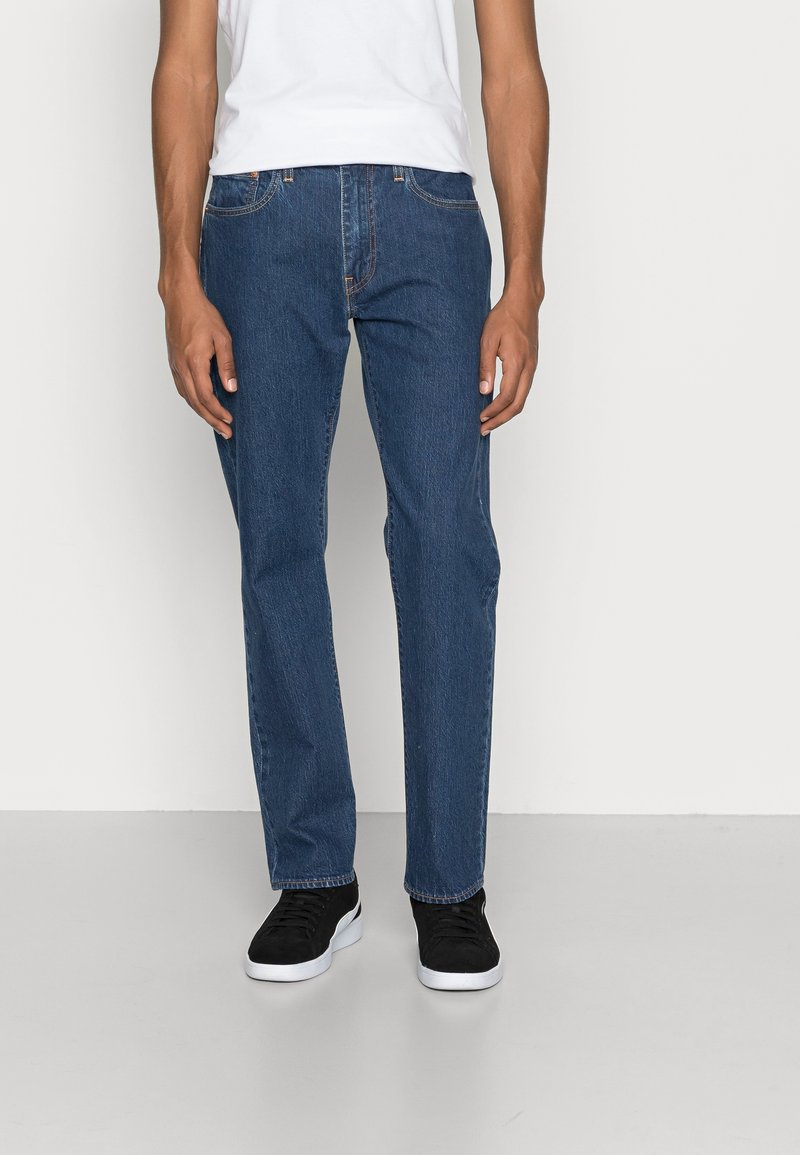 Hombre vistiendo jeans ajustados de color azul, zapatillas negras con suelas blancas y una camiseta sin mangas blanca frente a un fondo liso.