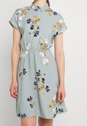 Jasnoniebieska sukienka z krótkim rękawem, zapięta na guziki przy kołnierzyku, z kwiatowym wzorem w kolorach białym, czarnym i złotym, noszona przez kobietę ze spuszczonymi rękami.