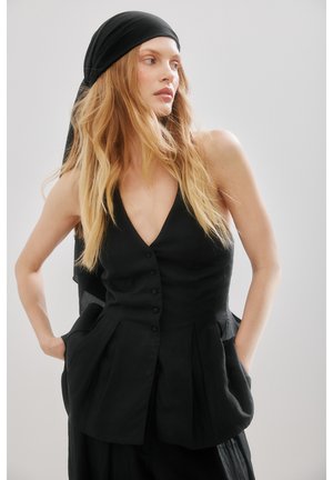 Kobieta o długich blond włosach, ubrana w czarną bezrękawnik zapinany na guziki i chustkę na głowie, patrząca na bok z rękami w kieszeniach.