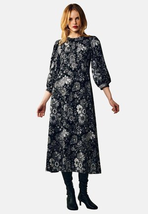 Lång svart klänning med blommönster i grått och vitt, med puffärmar och figurnära överdel. Stylad med svarta knähöga stövlar.