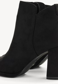 Bottines noires en daim avec un talon bloc haut et un bord supérieur courbé, présentées sur un fond blanc uni.