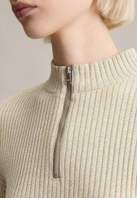 Hellbeige geripptes Sweatshirt mit hohem Kragen und Halbzipper. Der Stoff hat eine weiche Textur und ein dezentes Muster.