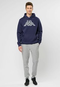 Sudadera con capucha de color azul marino con un gran logo blanco, combinada con pantalones jogger grises. La sudadera tiene un bolsillo frontal y capucha con cordón.