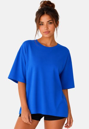 Jeune femme aux cheveux bruns en chignon, portant un t-shirt ample bleu vif et un short noir sur un fond clair uni.