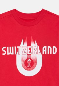 T-shirt in cotone rosso con la scritta "SVIZZERA" in lettere bianche in grassetto, con un logo circolare e accenti grafici al centro.