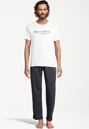 Mand står barfodet iført hvid Marc O’Polo T-shirt og sorte løse bukser foran en ensfarvet lys baggrund.