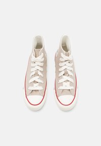 Béžové vysoké tenisky s bílými tkaničkami, gumovou špičkou, červeným pruhem na mezipodešvi a logovými nášivkami po stranách.