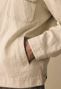 Jachetă texturată de un bej deschis, cu buzunare pe piept și deschideri laterale pentru mâini, având un design curat și un detaliu de buton contrastant la tiv.