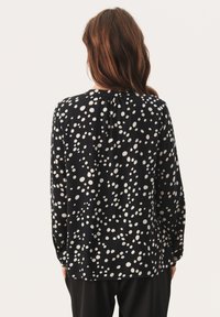 Vrouw met lang bruin haar, gekleed in een zwarte blouse met onregelmatige witte stippen, weergegeven van achteren tegen een effen achtergrond.