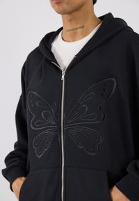 Μαύρο φούτερ με φερμουάρ από βαμβάκι, διαθέτει υφαντή κεντημένη πεταλούδα μπροστά. Περιλαμβάνει τσέπη καγκουρό και άνετη κουκούλα.