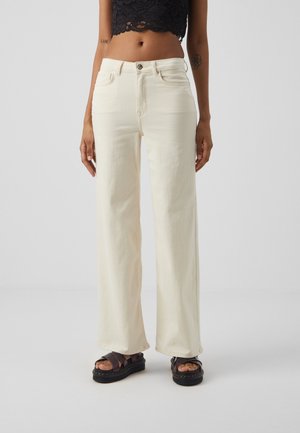 Jean large couleur crème avec poches avant, fermeture à boutons argentés et texture lisse, associés à des sandales à plateforme foncées.