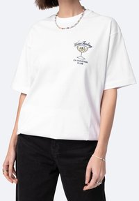 Biała bawełniana koszulka z krótkim rękawem, z nadrukowanym graficznym wzorem kieliszka wina oraz napisem "Degustacja Wina W KLUBIE WAKACYJNYM" w kolorze czarnym.